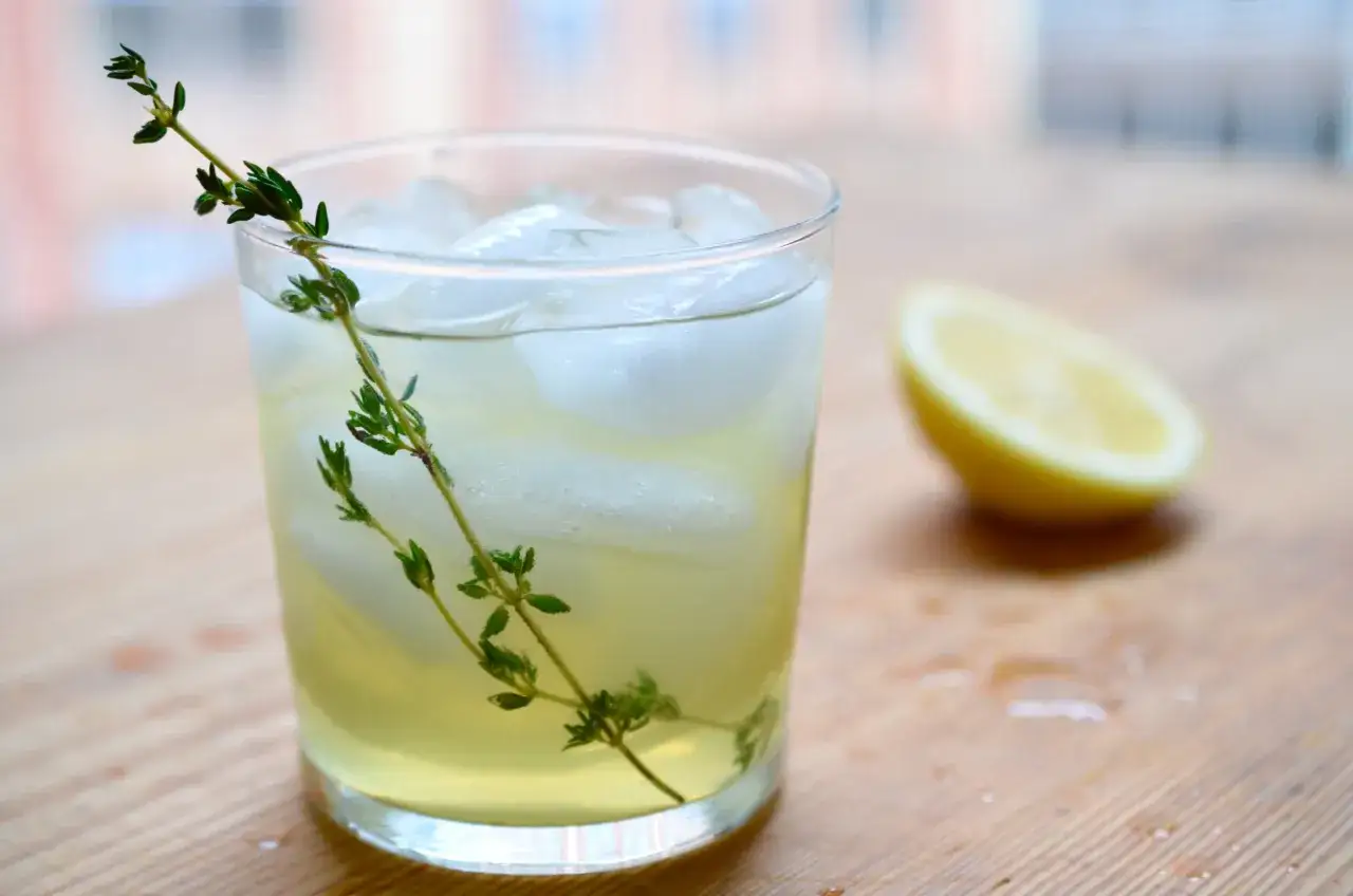 Ett glas med is och en ljusgul dryck, garnerad med en kvist timjan. En halv citron ligger i bakgrunden. En perfekt limoncello spritz f&ouml;r en solig dag.