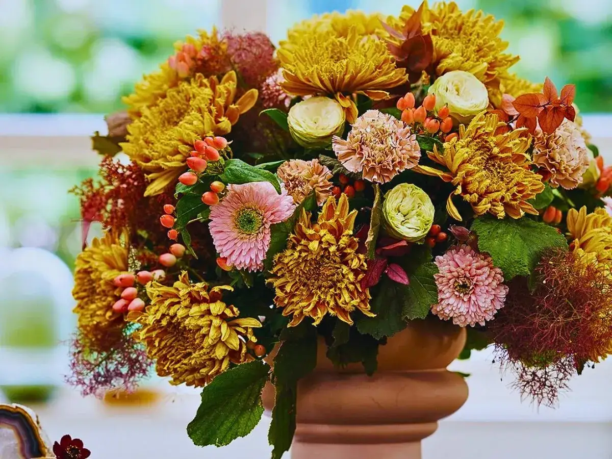 Magnifique composition automnale de chrysanthèmes jaunes, roses pâles, baies orangées et feuillage dans un vase en terre cuite.