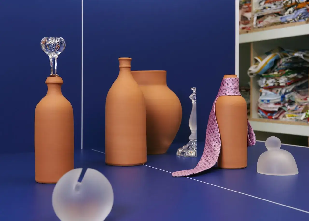 Un articolo di Hermès: una cravatta rosa su una bottiglia di terracotta, con vasi e oggetti in vetro su sfondo blu.