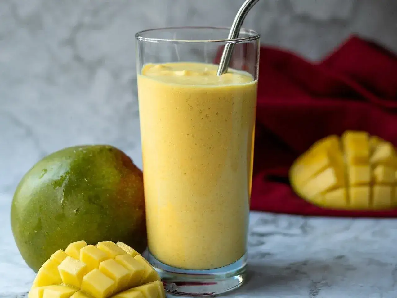 Orzeźwiający mango lassi w szklance, obok świeże mango i kawałki owocu. Idealny napój na upalne dni.