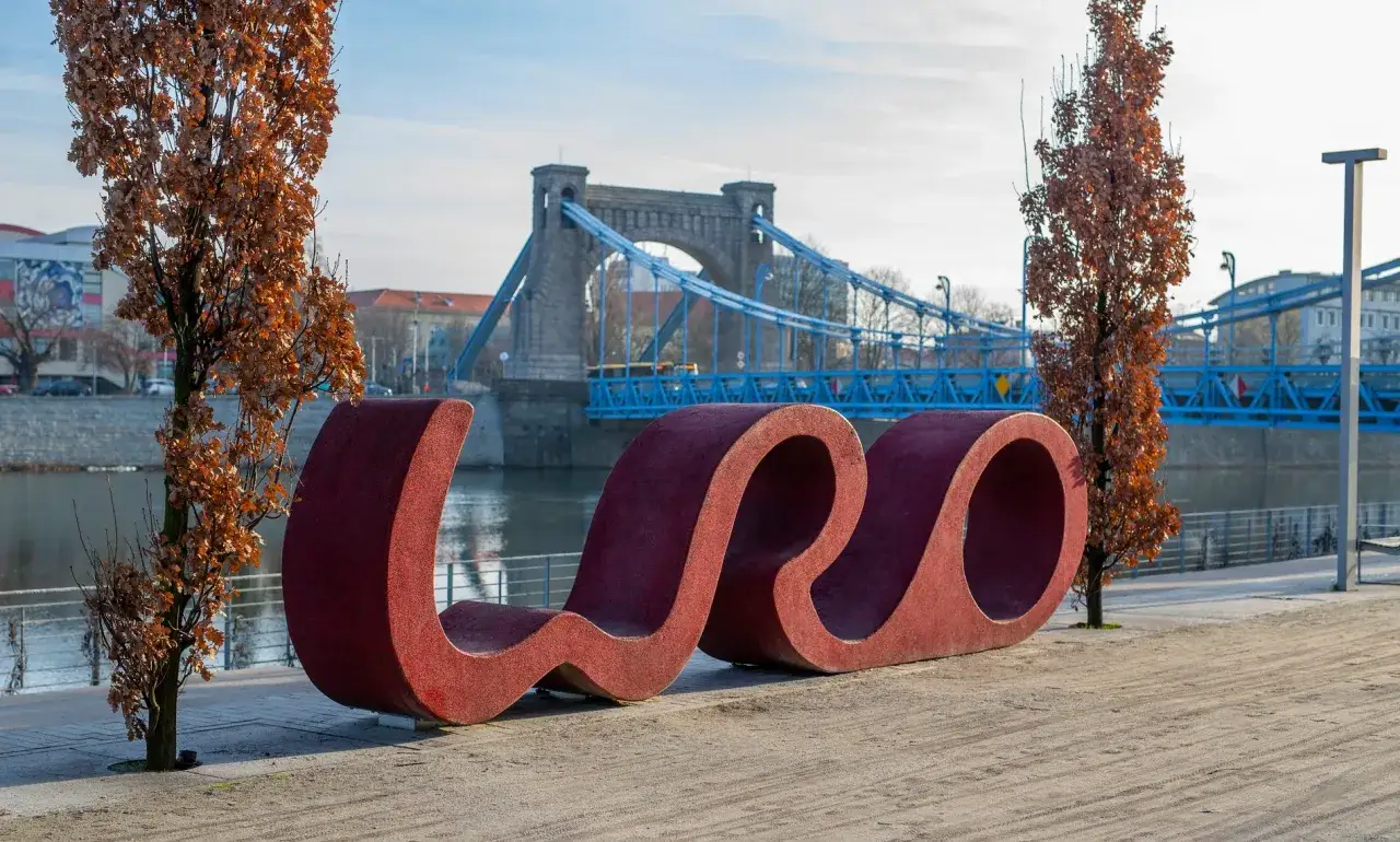 Czerwona, falista rzeźba na brzegu Odry, obok drzew z brązowymi liśćmi. W tle błękitny most i zabytkowa architektura – nietypowe atrakcje Wrocławia.
