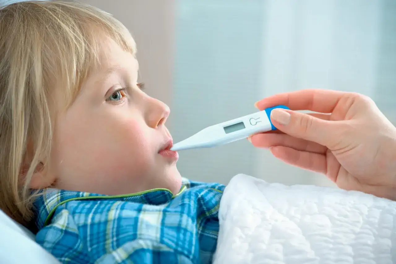 Dziecko z gorączką leży w łóżku, a ręka mamy mierzy mu temperaturę termometrem.
