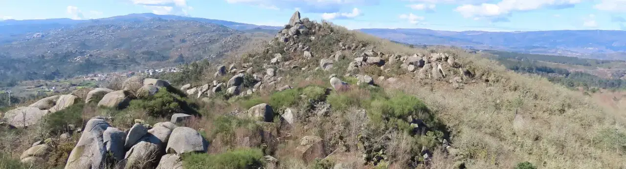 Paisaje rocoso con vegetaci&oacute;n baja y un pueblo a lo lejos. La **pena corneira** se alza imponente entre las rocas.