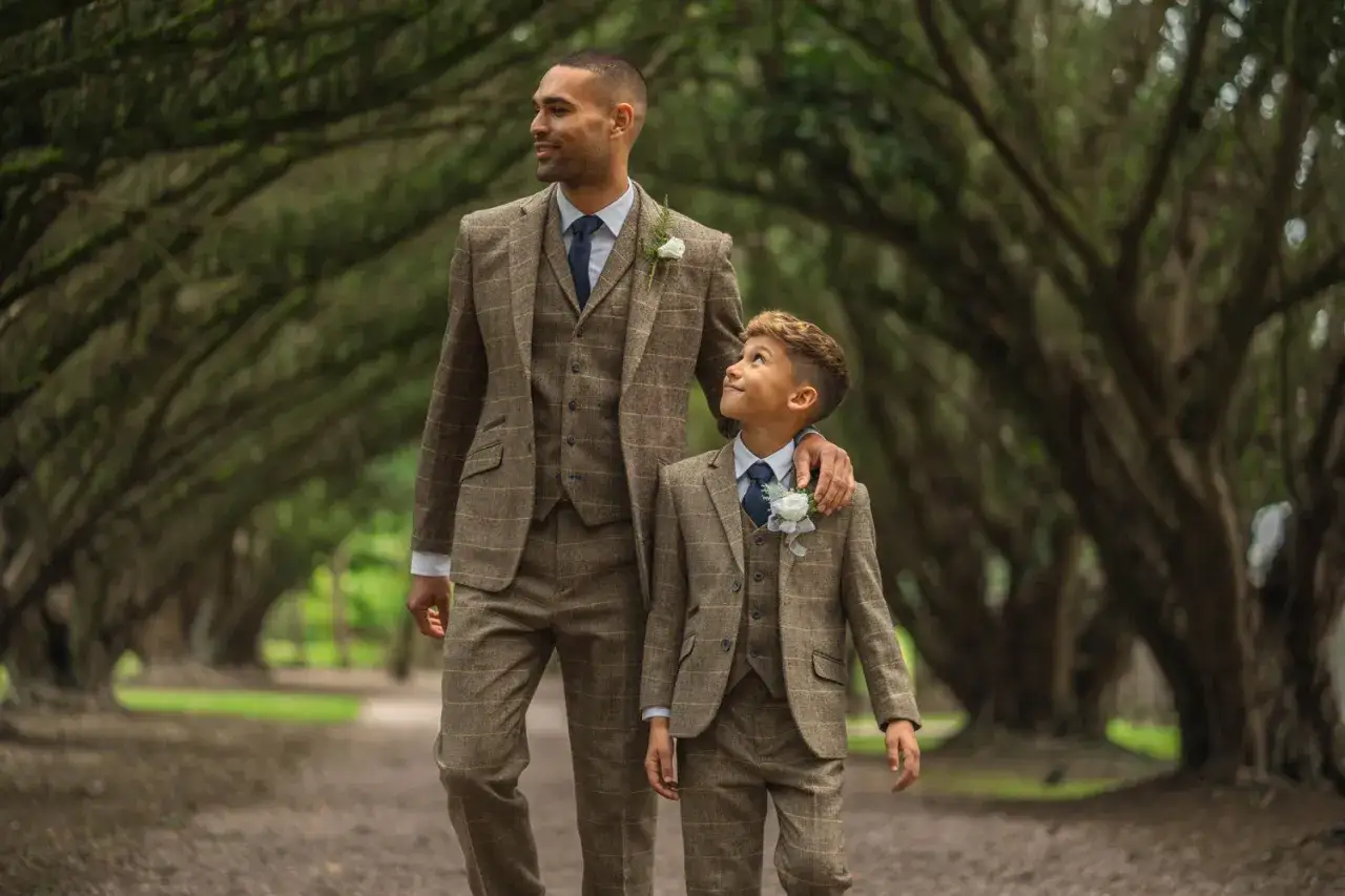 A man and boy in matching tweed suits, perfect for dressy casual attire at a wedding, walk under a canopy of trees.