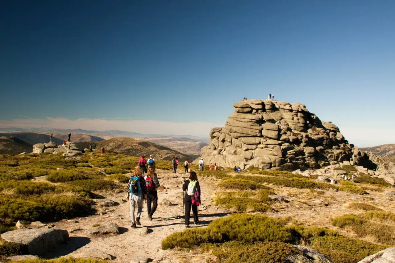Senderistas disfrutan de las vistas de los picos de la Sierra de Guadarrama, con formaciones rocosas y vegetación baja bajo un cielo azul.