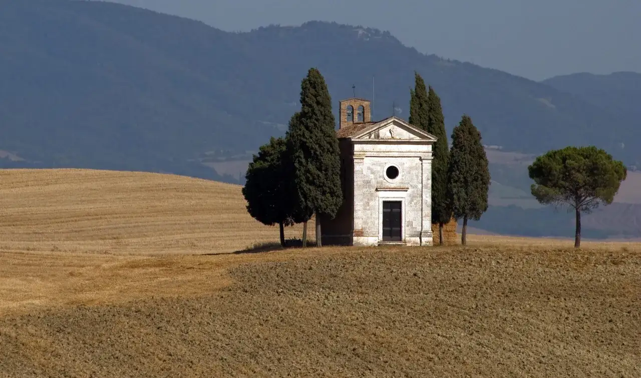 Cappella della Madonna di Vitaleta in Val d'Orcia, circondata da cipressi e campi dorati, con dolci colline sullo sfondo.