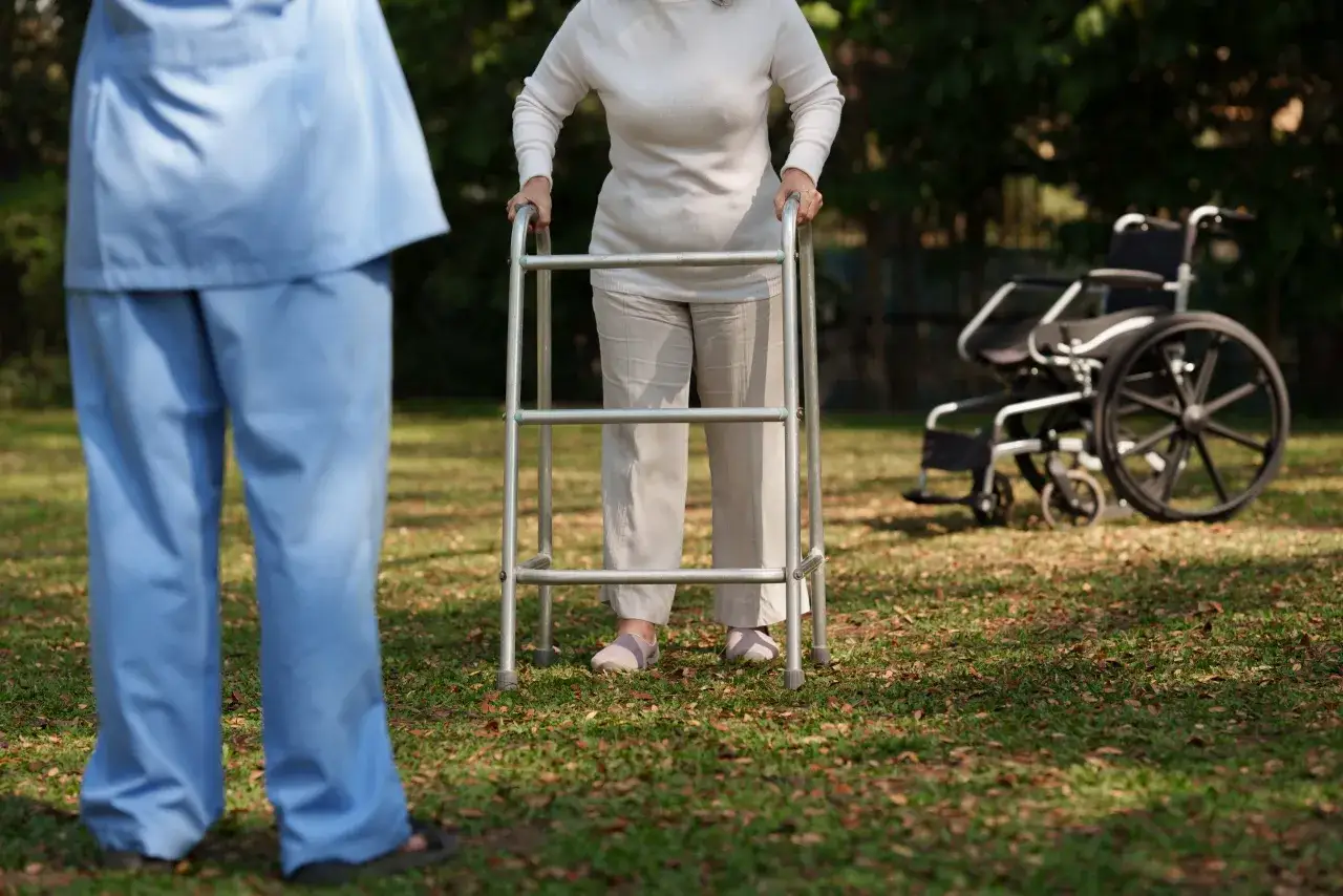 Assistente supporta una persona con **alzheimer e difficoltà a camminare**, che usa un deambulatore. Sfondo con sedia a rotelle.