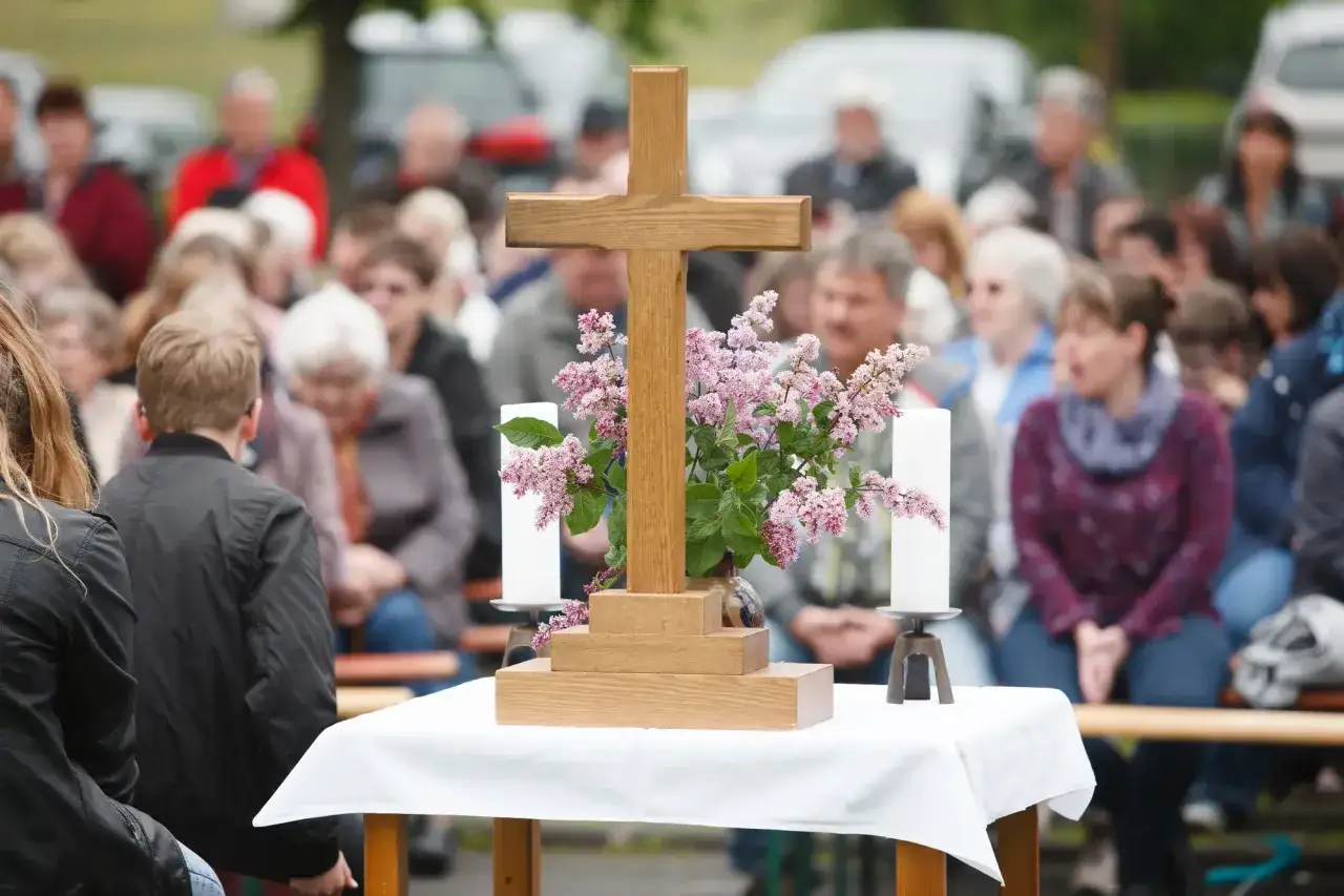 Holzkreuz mit Blumen und Kerzen vor einer versammelten Gemeinde. Ein Symbol für Glauben und Gemeinschaft, vielleicht im Kontext der Kirchensteuer Bayern.