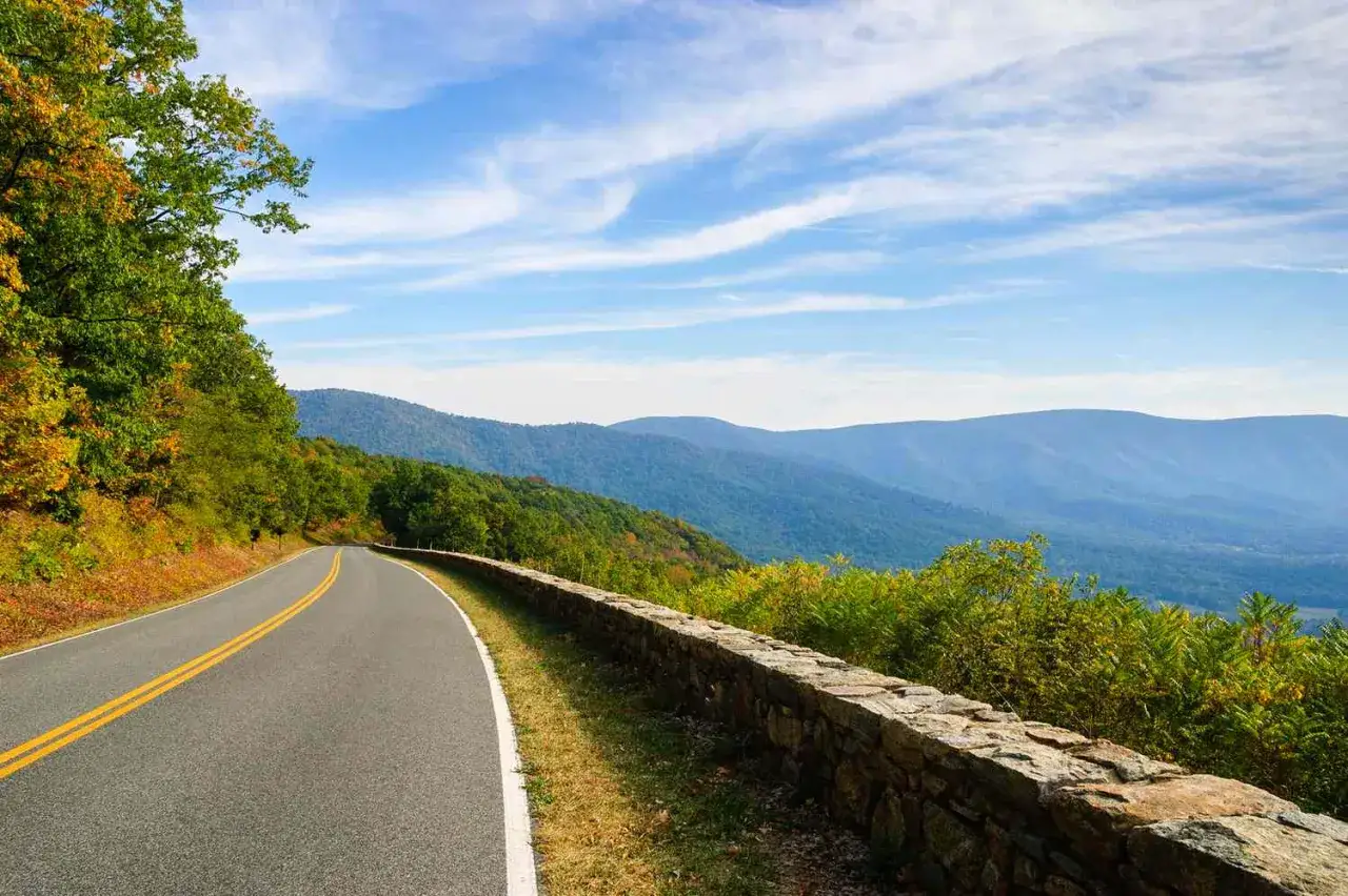 Scenic mountain road, perfect for weekend trips from Richmond, VA. Lush green trees line the winding asphalt, leading to breathtaking blue mountain vistas under a cloudy sky.