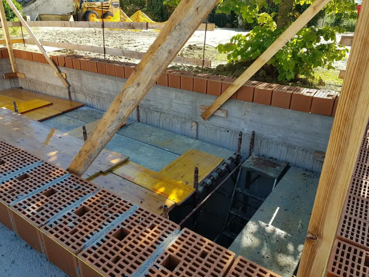 Costruzione in corso: blocchi di laterizio termico pronti per essere posati su fondamenta in cemento, con impalcature in legno e un escavatore sullo sfondo.