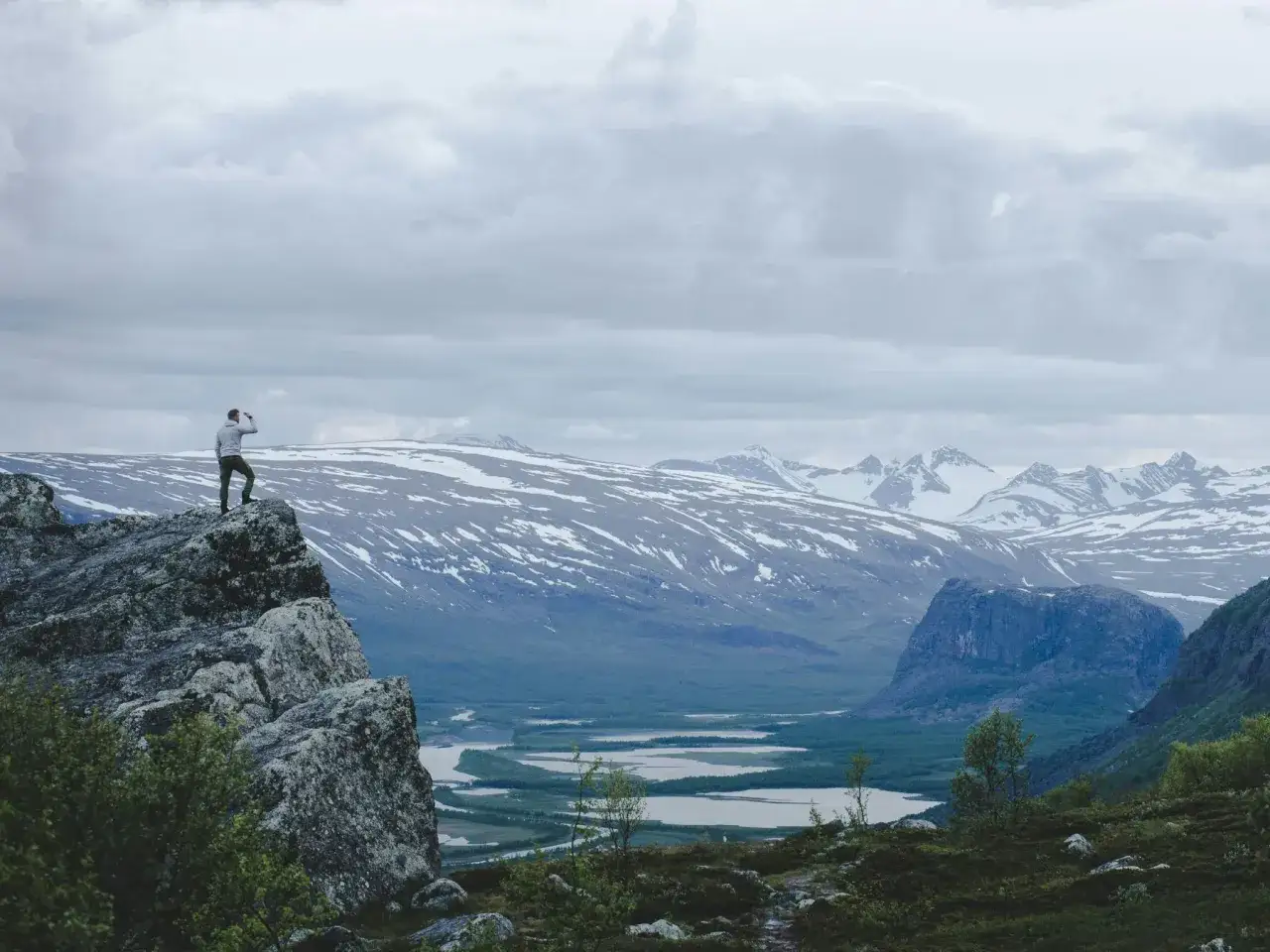 Vandrare blickar ut &ouml;ver fj&auml;llandskap med sn&ouml;kl&auml;dda toppar, en sj&ouml; och en m&auml;ktig klippa. En vy som p&aring;minner om Saltoluokta och Kvikkjokk.