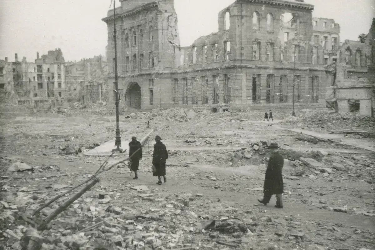 Zniszczone budynki po wojnie, symbolizujące potrzebę reparacji wojennych od Niemiec. Ludzie przechodzą przez gruzy.