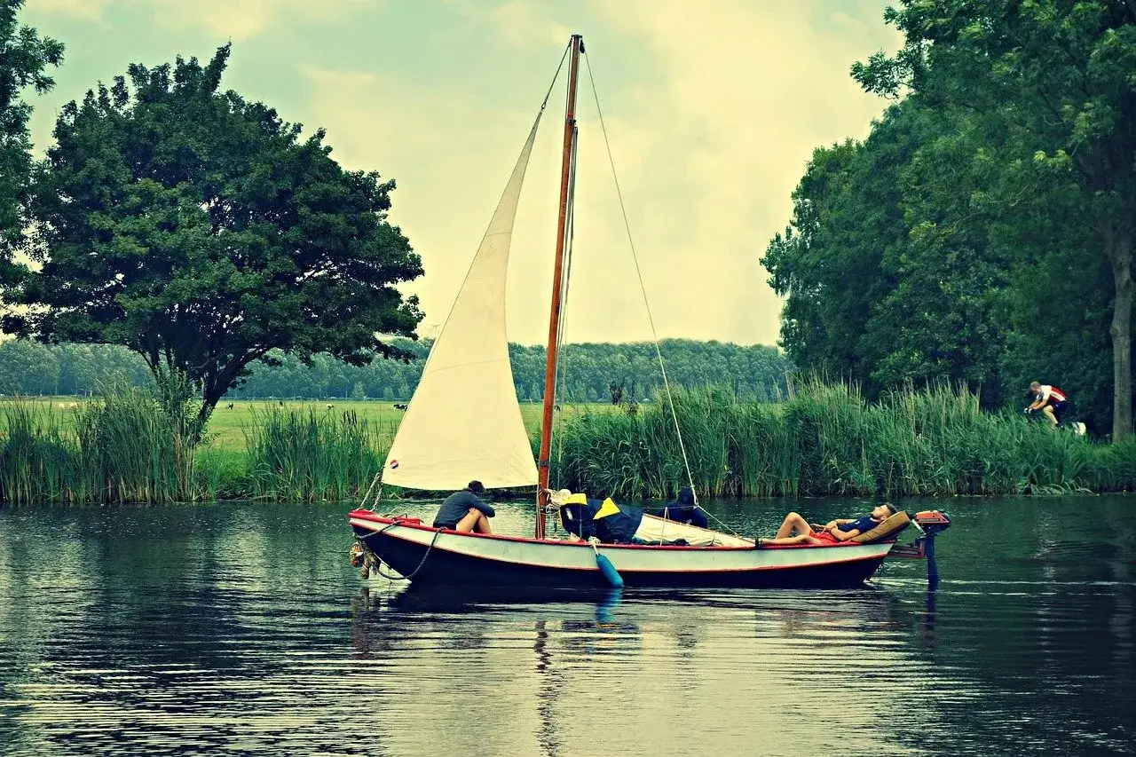 Un bateau &agrave; voile navigue sur un canal bord&eacute; de verdure. Des personnes se d&eacute;tendent &agrave; bord, tandis qu'un cycliste passe sur la rive.