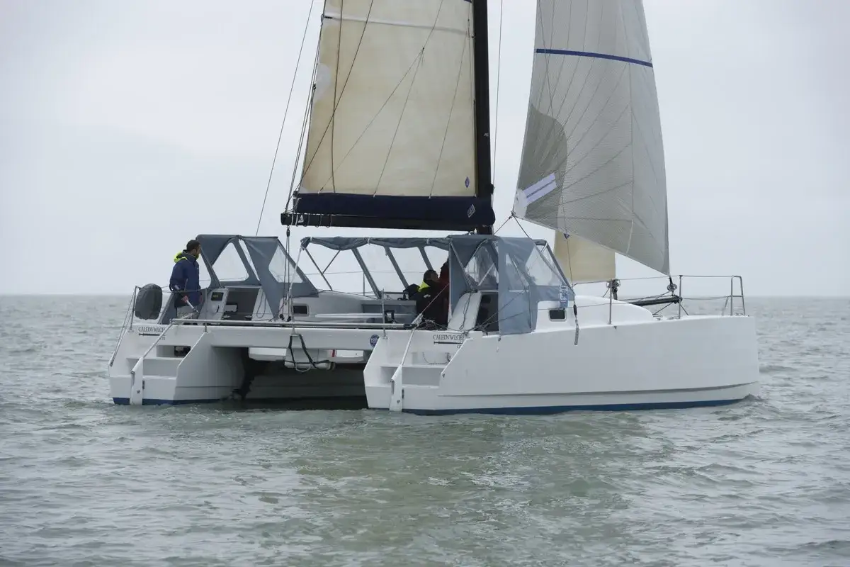 Un catamaran blanc navigue sur une mer grise. Deux personnes sont à bord de cette bicoque bateau, profitant de la voile.