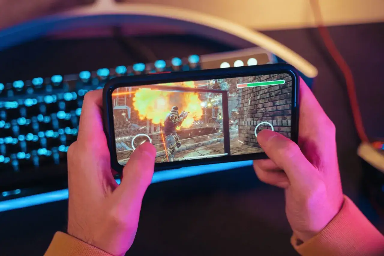 Händerna håller en mobil med ett actionfyllt spel. En explosion syns på skärmen, perfekt för roliga spel på mobilen.