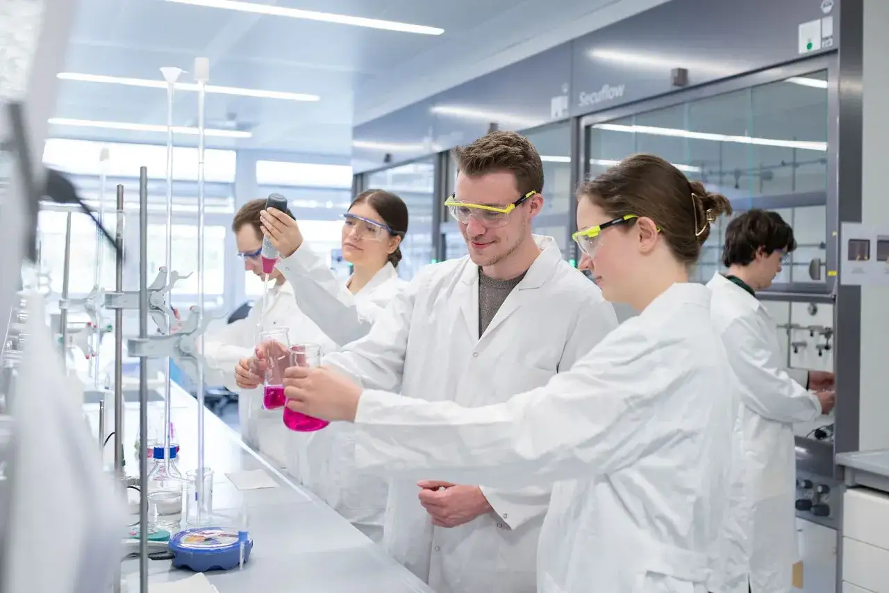 Studenten im Chemietechnik Studium f&uuml;hren Experimente durch. Sie tragen Laborkittel und Schutzbrillen und arbeiten mit Reagenzgl&auml;sern und Pipetten.