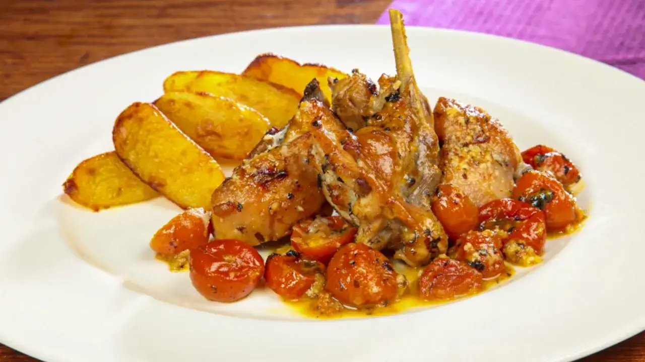 Un saporito coniglio al forno, dorato e succulento, servito con patate arrosto e pomodorini confit su un piatto bianco.