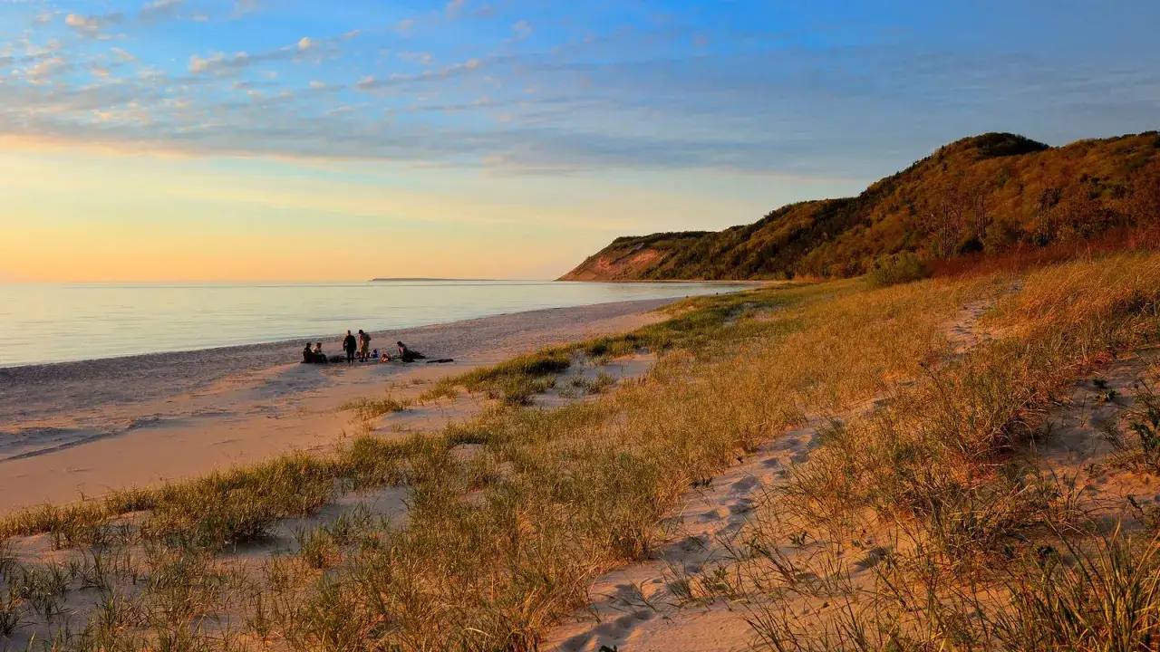 Zachód słońca na piaszczystej plaży, z łagodnymi falami i zalesionym wzgórzem w tle. Piękny krajobraz, jak z **parków narodowych w Stanach Zjednoczonych**.