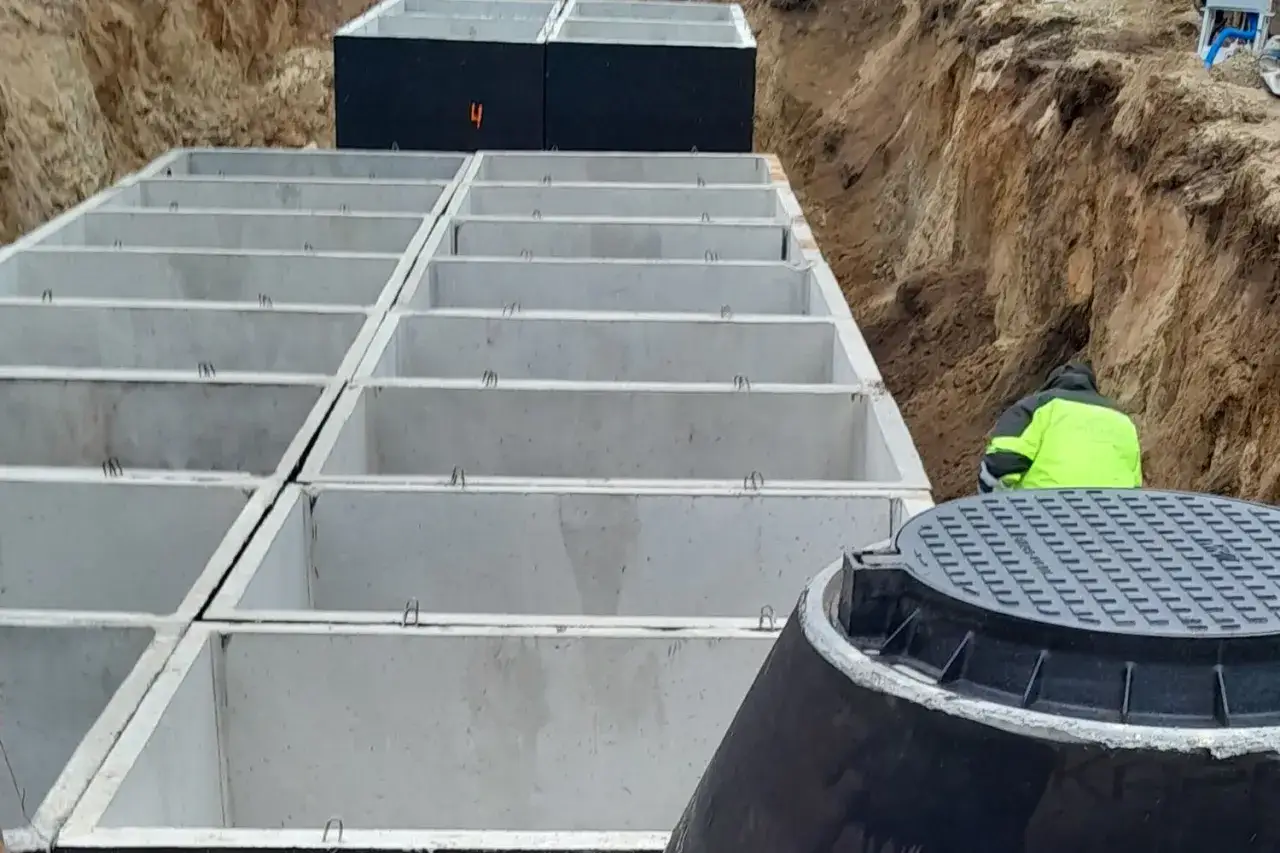 Budowa zbiornika przeciwpożarowego. Betonowe moduły ułożone w wykopie, pracownik w kamizelce odblaskowej.