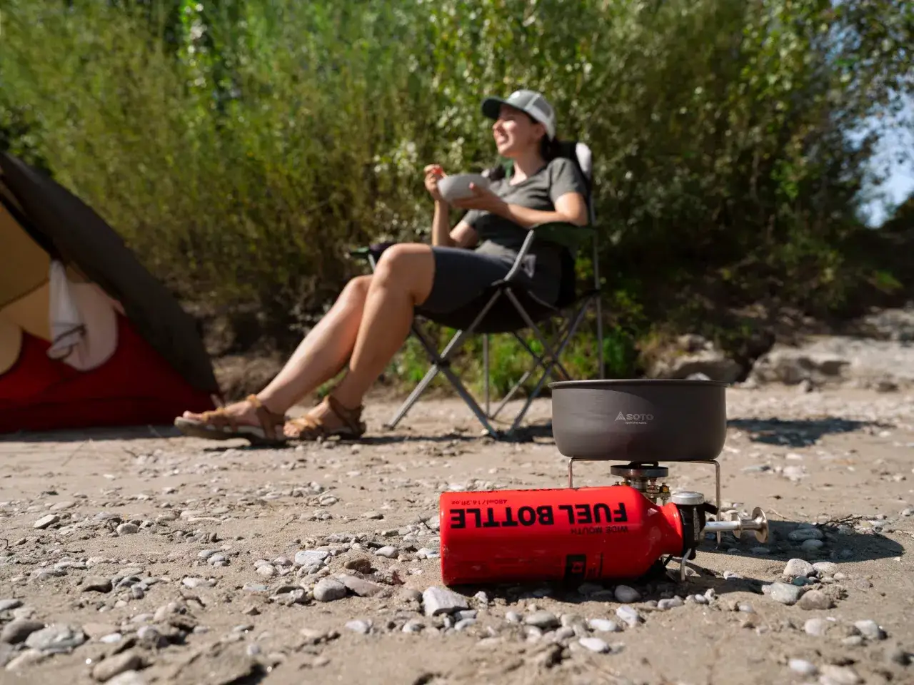 Soto Stormbreaker Kocher mit Brennstoffflasche am Strand. Eine Frau sitzt im Hintergrund in einem Campingstuhl.