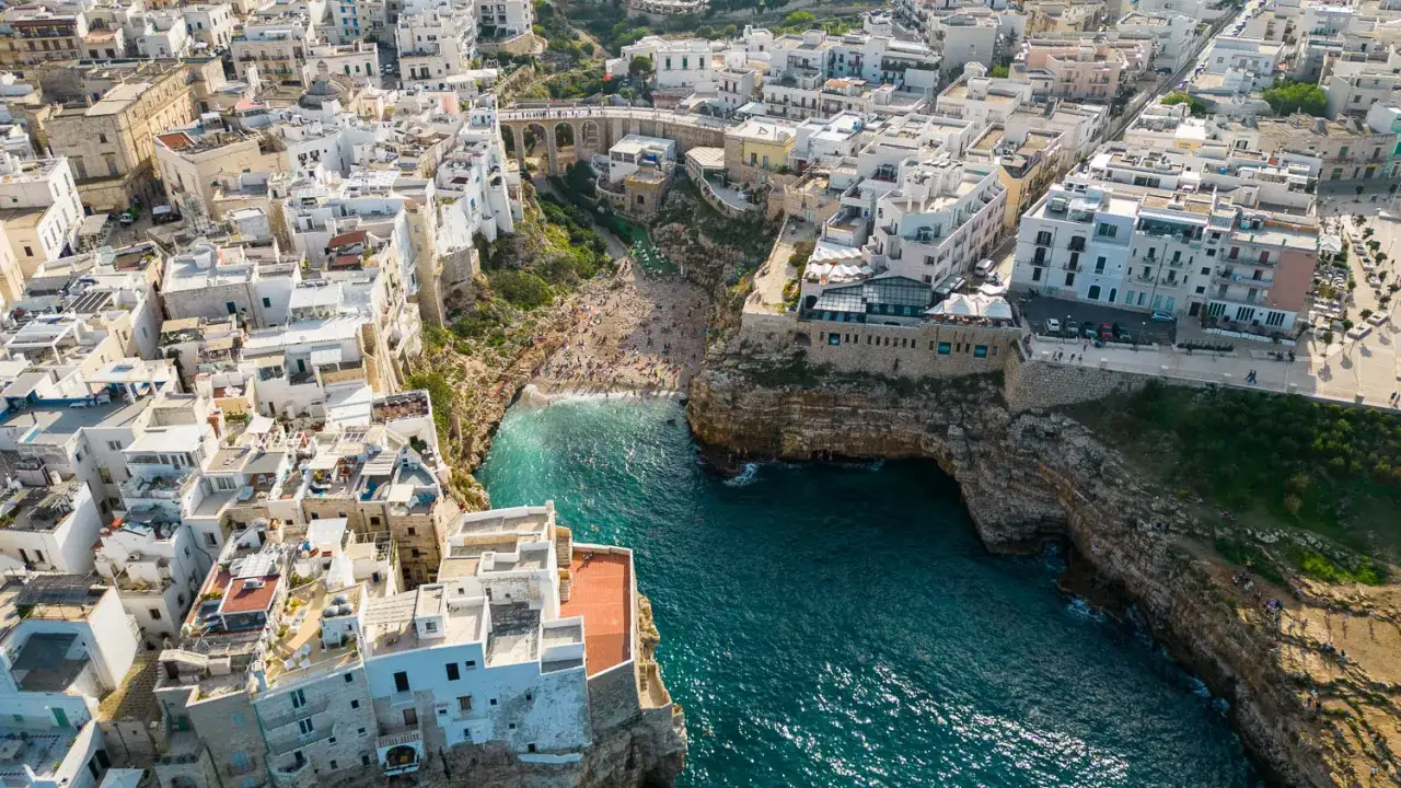 Vue aérienne de Polignano a Mare, avec sa célèbre plage nichée entre les falaises. Les maisons blanches s'accrochent aux rochers, surplombant les eaux turquoise.