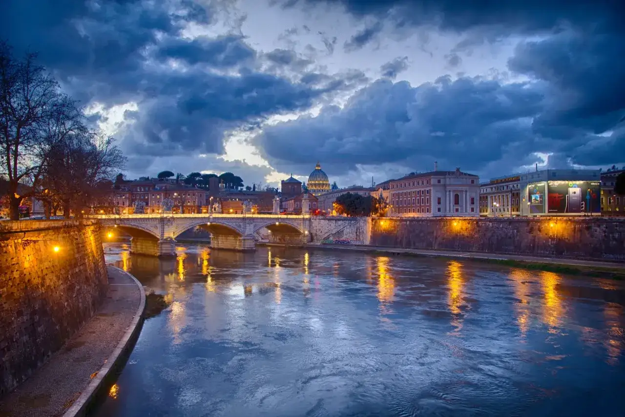Il Tevere riflette le luci di Roma al tramonto, con la cupola di San Pietro sullo sfondo. Un'atmosfera magica che anticipa la vivace **Trastevere nightlife**.