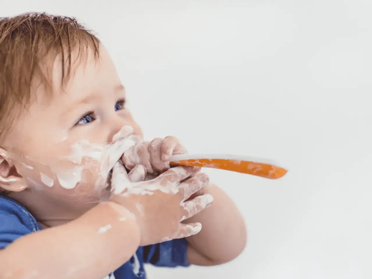Kleiner **Joghurt Baby** mit Joghurt im Gesicht und auf den H&auml;nden, der einen L&ouml;ffel h&auml;lt.