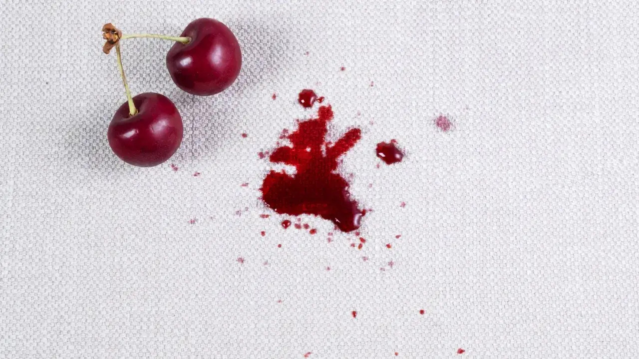 Dwie wiśnie leżą obok rozlanej plamy z wiśni na jasnym materiale.