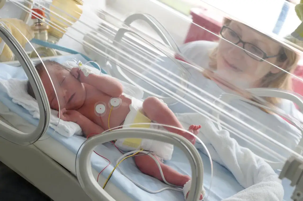 Un bebé prematuro duerme en una incubadora de bebés, con cables conectados y una enfermera observando con ternura.