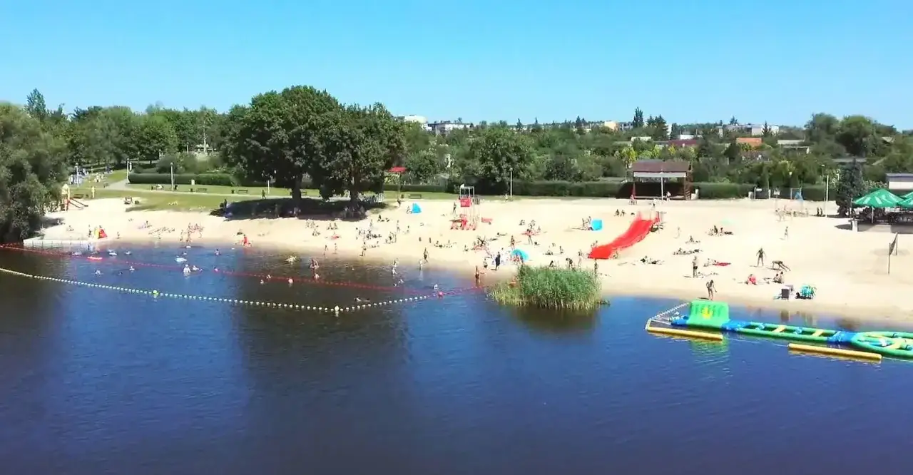 Słoneczny dzień nad jeziorem Grzymisławskim. Ludzie odpoczywają na plaży, kąpią się i bawią na dmuchanych atrakcjach.