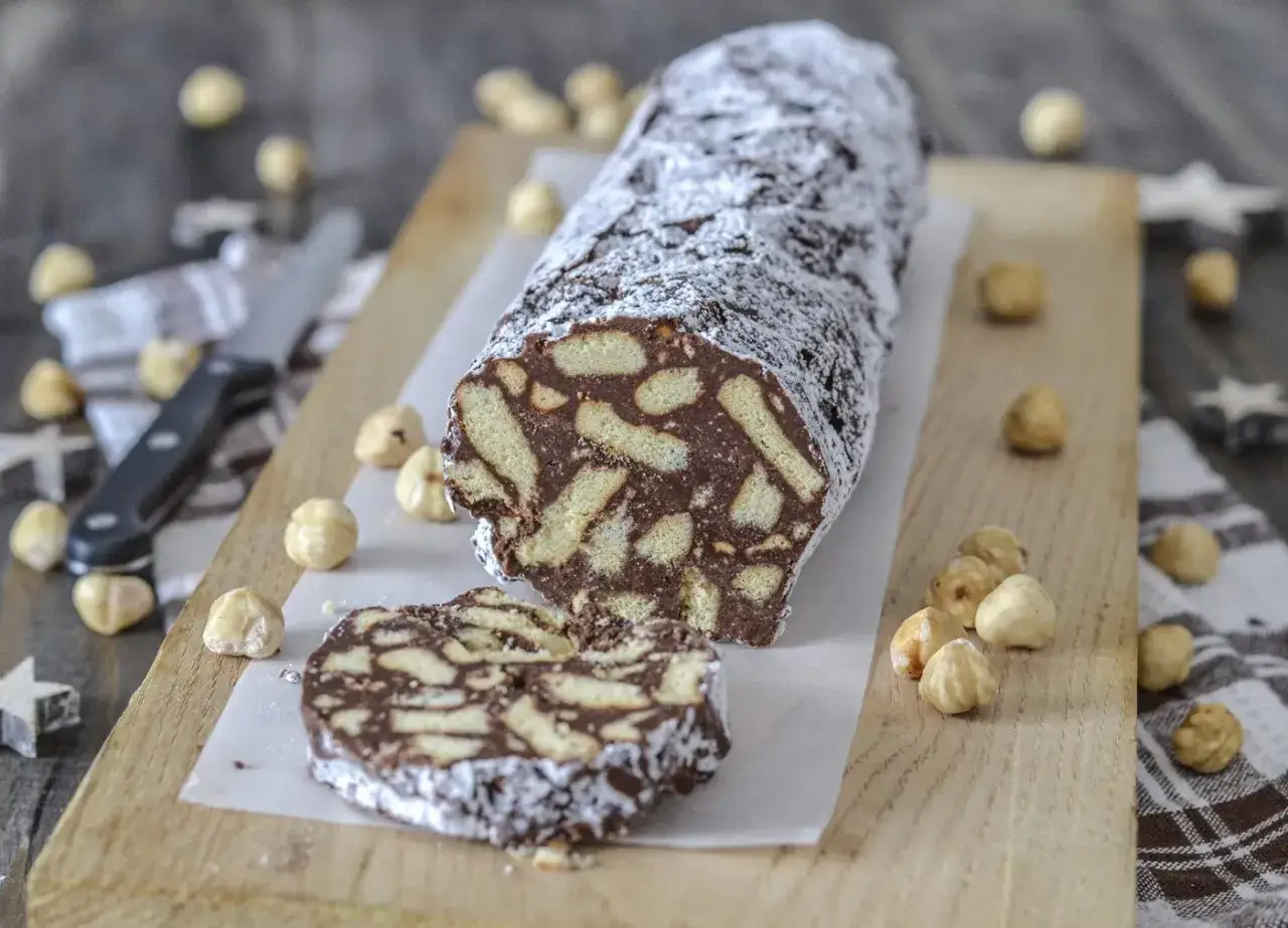 Un delizioso salame al cioccolato, cosparso di zucchero a velo, con nocciole sparse attorno.