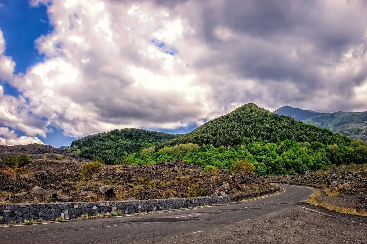 Strada tortuosa tra rocce laviche e boschi lussureggianti, ideale per itinerari moto Sicilia. Nuvole drammatiche nel cielo.