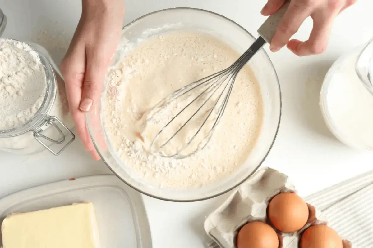 Ręce mieszają trzepaczką ciasto na pancake w szklanej misce. Obok mąka, jajka i masło.