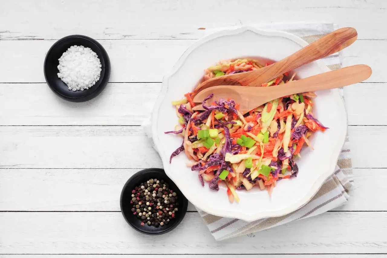 Deliciosa ensalada de col con zanahoria rallada, repollo morado y cebolleta. Servida en plato blanco con cucharas de madera.