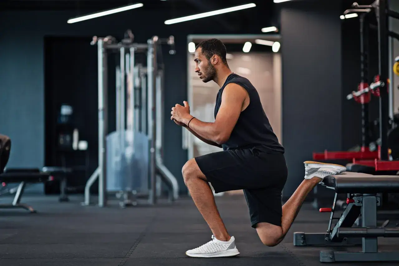 Mann macht einen ATG Split Squat im Fitnessstudio. Sein hinterer Fu&szlig; ruht auf einer Bank, w&auml;hrend er sich tief absenkt.