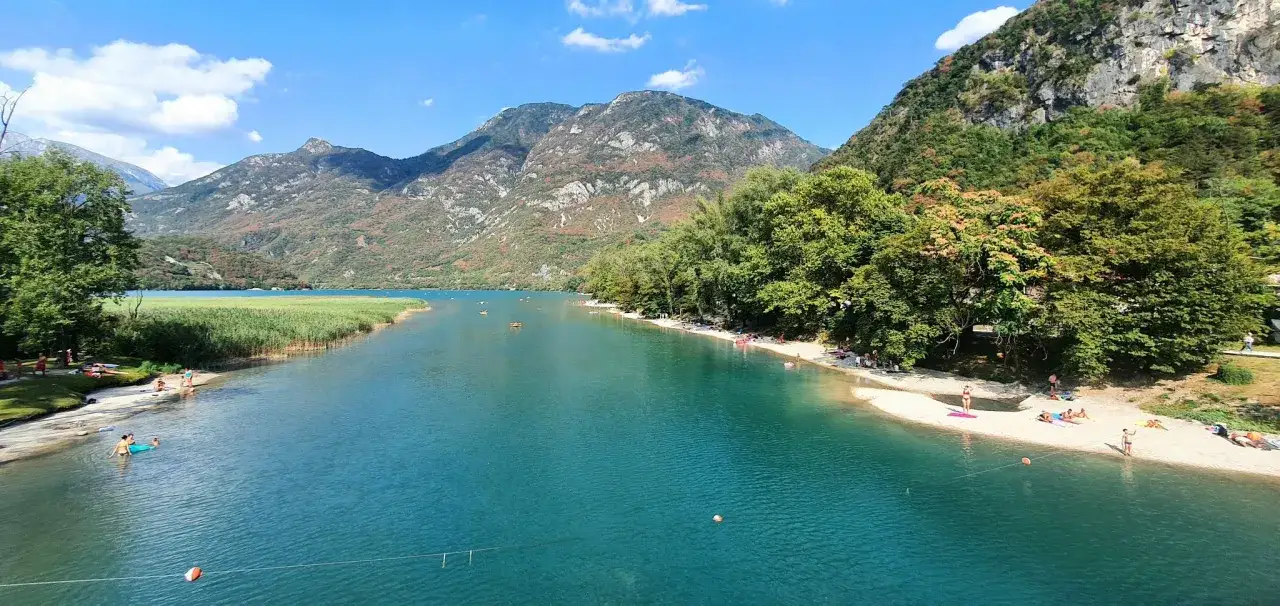 T&uuml;rkises Wasser am Lago di Cavazzo, umgeben von gr&uuml;nen H&uuml;geln und Bergen. Menschen entspannen am sandigen Ufer und genie&szlig;en das klare Wasser.