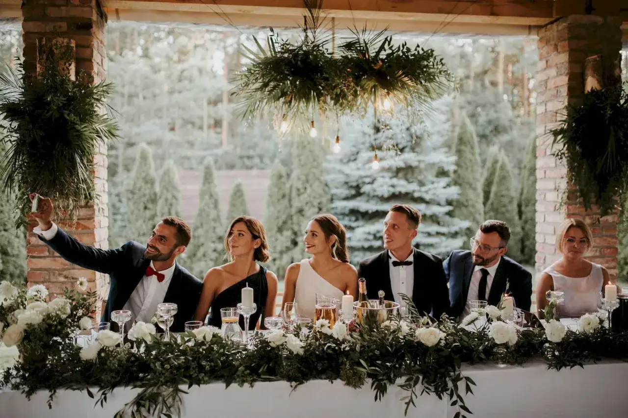 Goście weselni czekają na piosenki na wejście pary młodej na salę. Uśmiechnięty mężczyzna robi selfie.