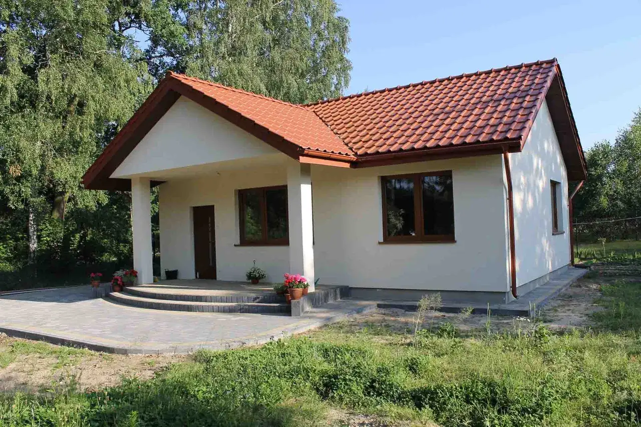 Nowy, biały dom z czerwonym dachem i brązowymi oknami, otoczony zielenią.
