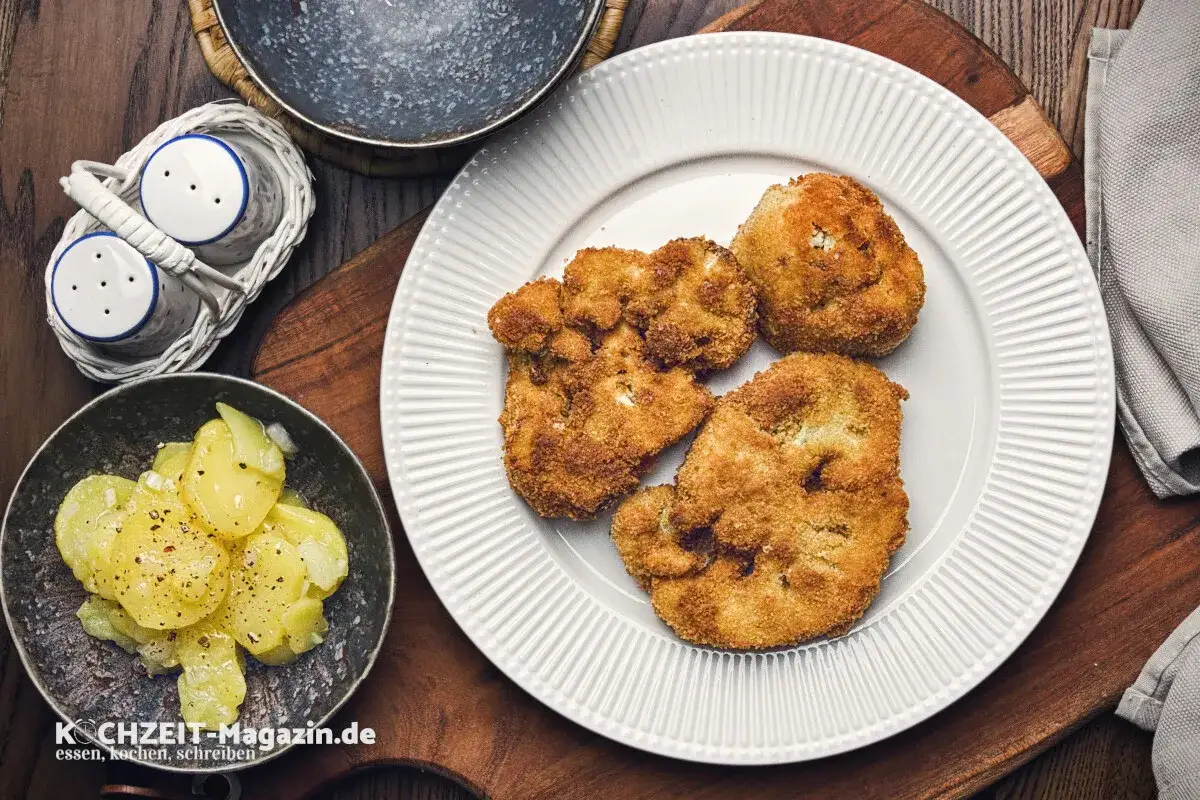 Knusprig panierter Blumenkohl auf einem wei&szlig;en Teller, daneben Kartoffelsalat und ein Salz- und Pfefferstreuer. Ein leckeres Gericht!