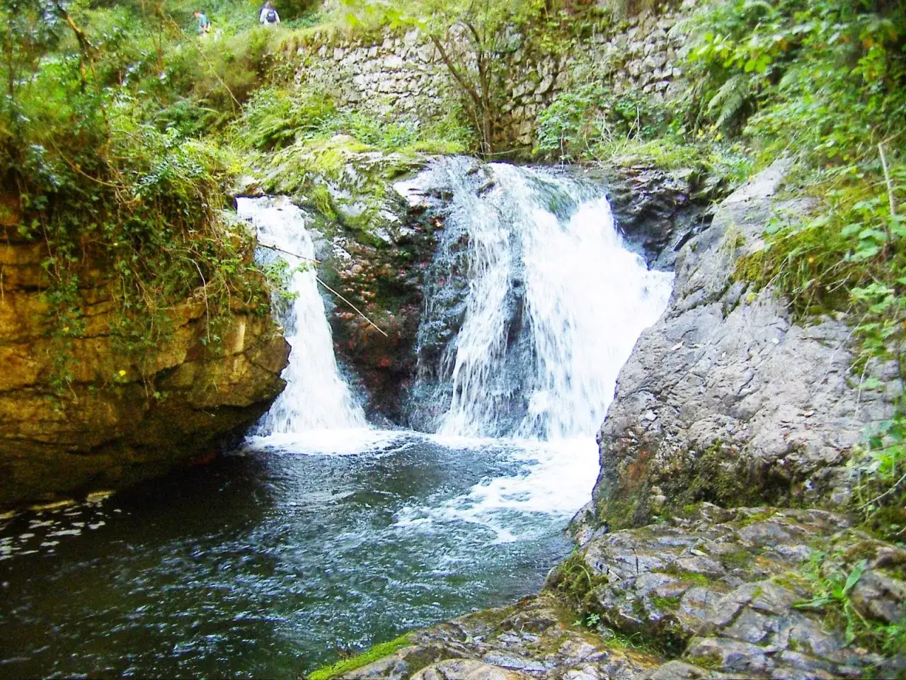 Cascada en la ruta del alba, con rocas cubiertas de musgo y vegetación exuberante. Dos personas caminan en la parte superior.