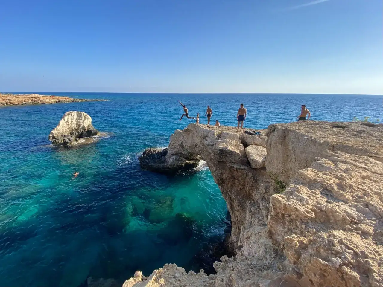 Ungdomar hoppar från klippor i det turkosa vattnet vid en naturlig stenbro. En perfekt dag för Ayia Napa aktiviteter.