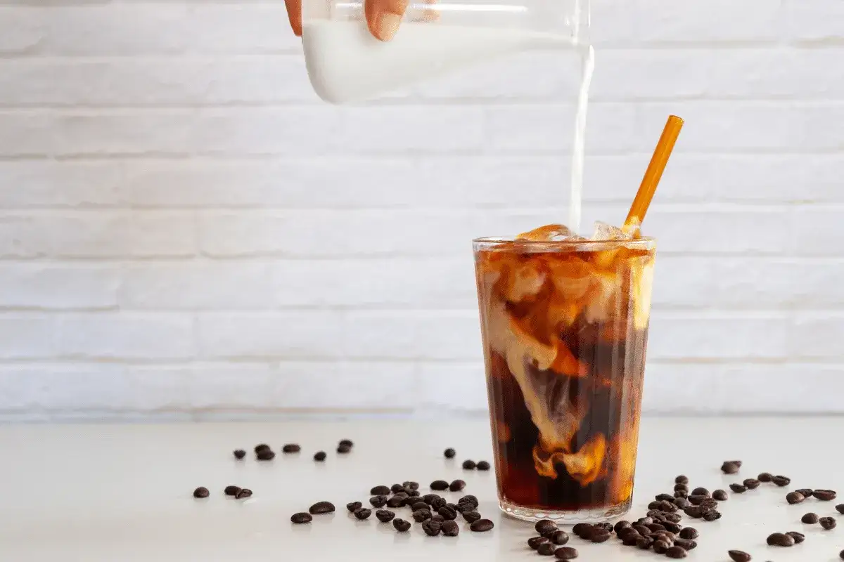 Milch flie&szlig;t in ein Glas mit Eis und Kaffee. Ein perfektes cold brew rezept f&uuml;r hei&szlig;e Tage. Kaffeebohnen liegen verstreut.