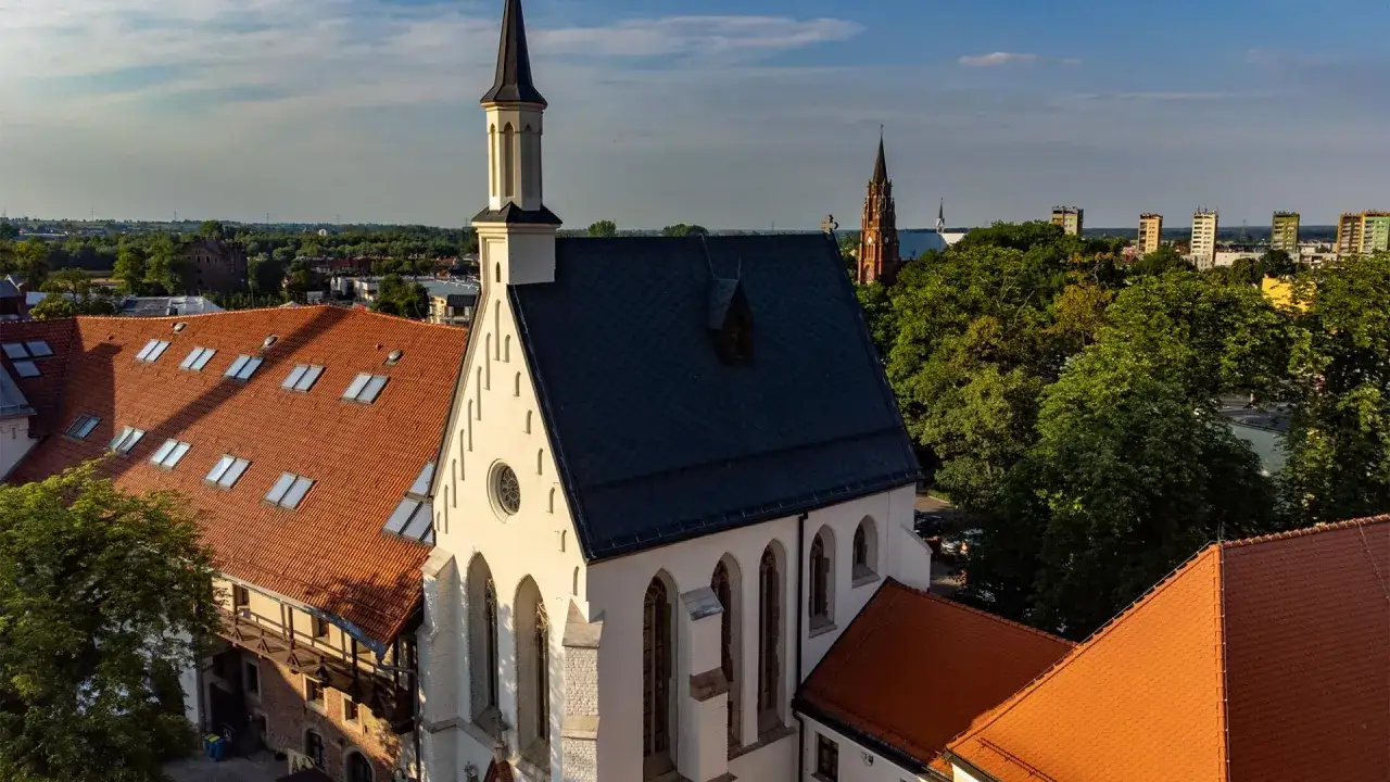 Widok z lotu ptaka na rynek w Raciborzu. Biały kościół z czarnym dachem i wieżą góruje nad ceglanymi dachami.