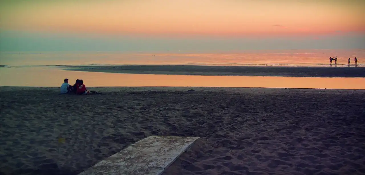 Un'alba a Rimini con persone che passeggiano sulla riva e una coppia seduta sulla sabbia, mentre il cielo si tinge di colori caldi.