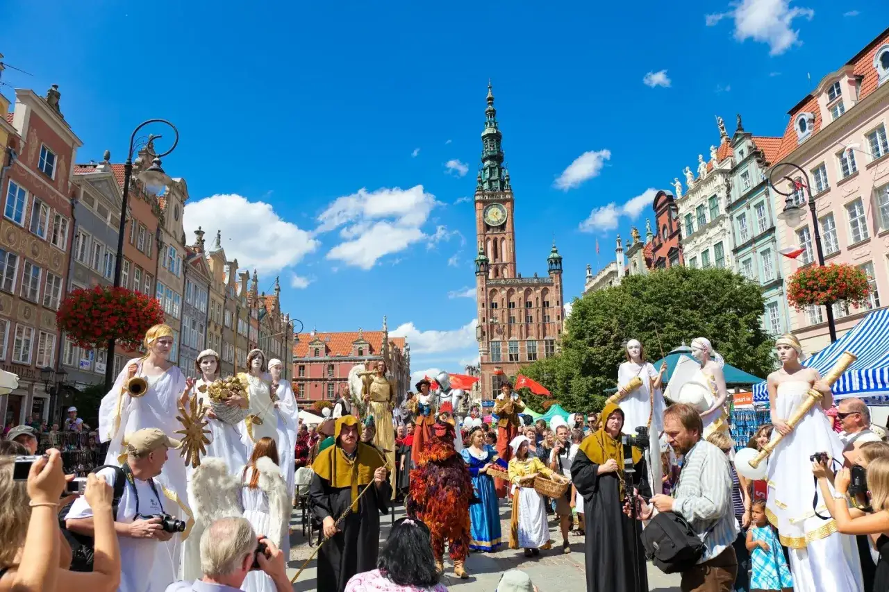 Kolorowy jarmark Gdańsk z postaciami na szczudłach i tłumem widz&oacute;w na tle zabytkowych kamienic.