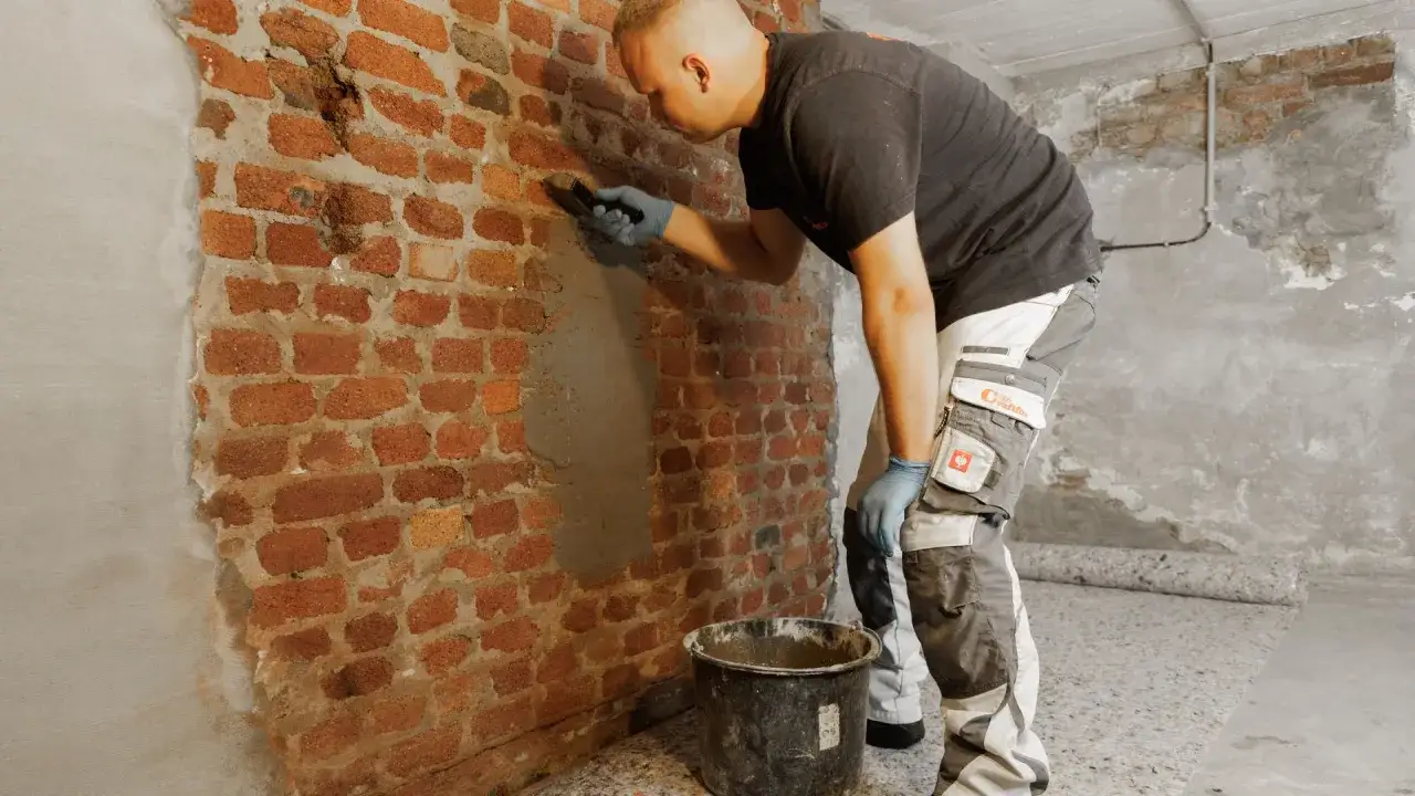 Mann repariert Ziegelwand, die nach Wasserschaden muffig riecht. Er tr&auml;gt M&ouml;rtel auf, um die besch&auml;digten Stellen zu versiegeln.