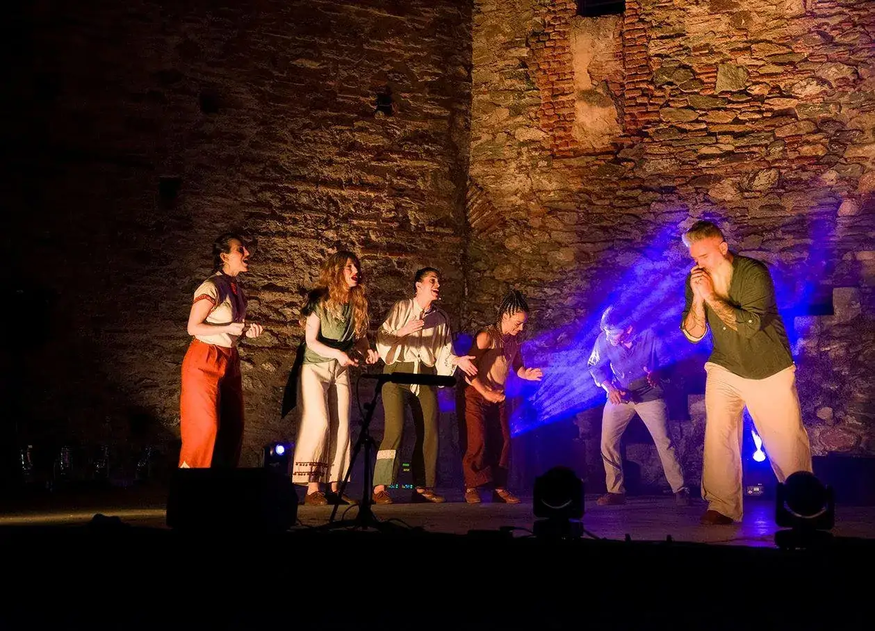 Sc&egrave;ne de th&eacute;&acirc;tre avec des acteurs sur fond de mur de pierre. Un homme barbu, peut-&ecirc;tre un percussionniste c&eacute;l&egrave;bre, chante avec passion.