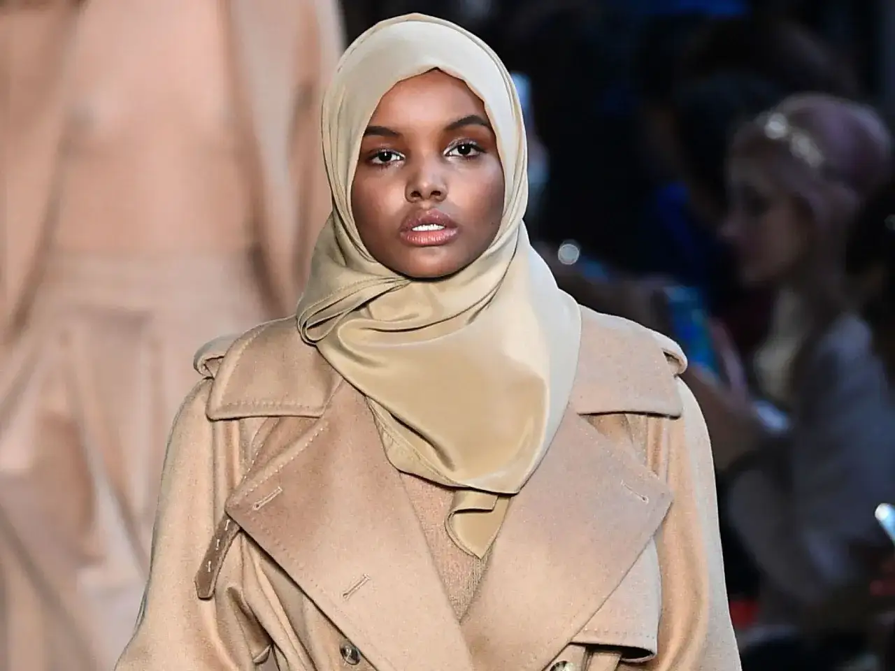 Halima Aden walks the runway in a tan trench coat and matching hijab, exuding confidence and style.