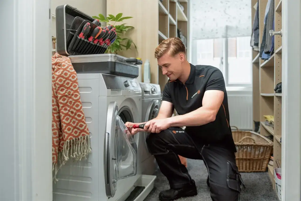 Monteur repareert een vastgelopen wasmachine. Een gereedschapskist en een deken liggen op de machine.