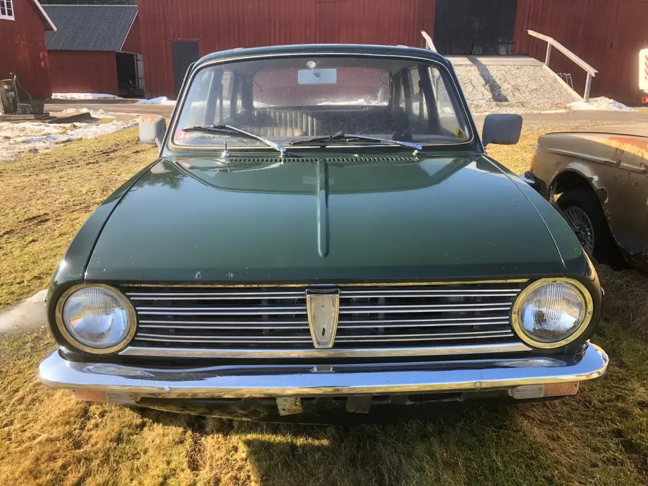 Gr&ouml;n Austin Maxi parkerad framf&ouml;r en r&ouml;d lada. Bilen har en kromad grill och runda str&aring;lkastare.